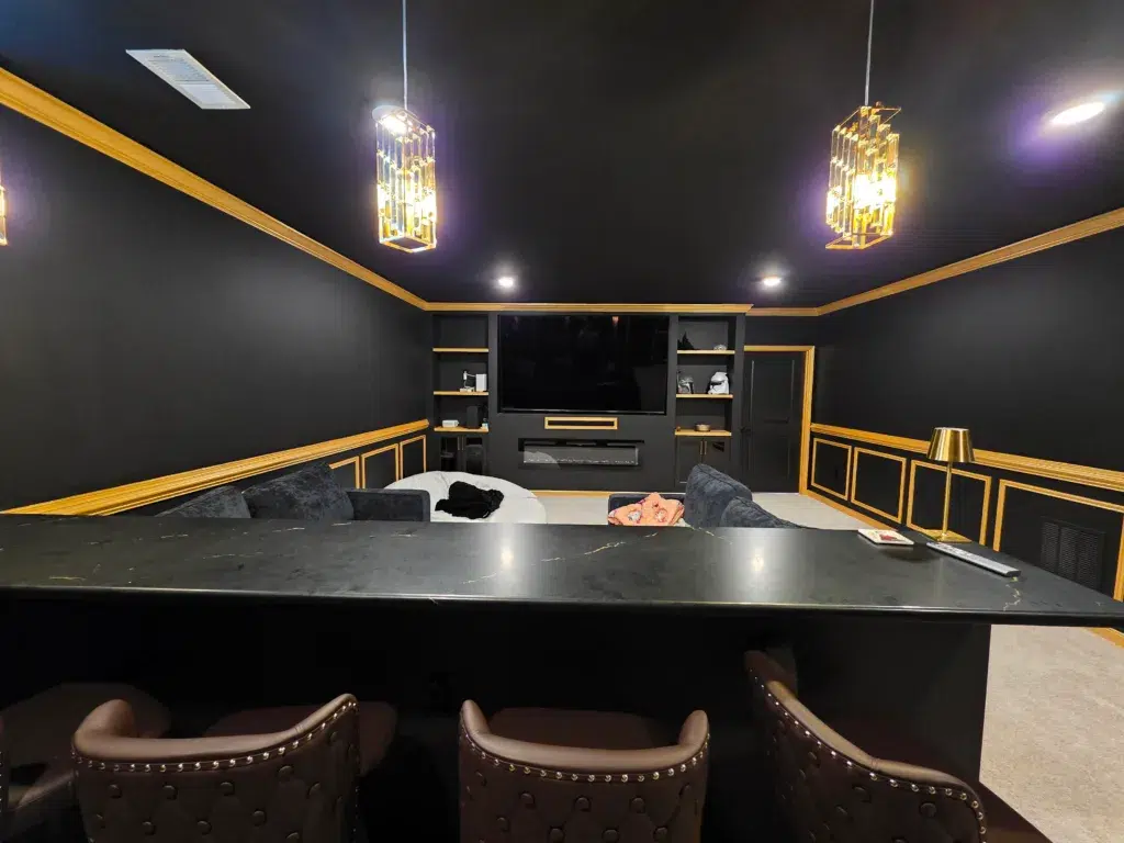 A professionally painted modern home theater with dark walls, ceiling, gold trim, and a sleek bar area.